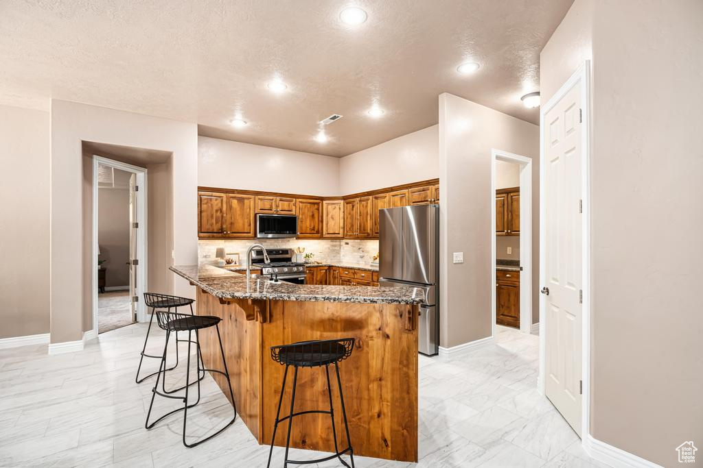 Kitchen with stainless steel appliances, brown cabinetry, a breakfast bar area, dark stone counters, and decorative backsplash