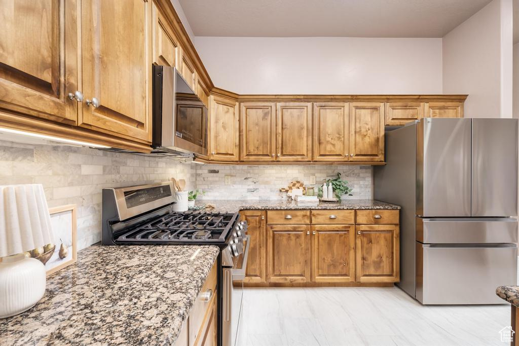 Kitchen featuring appliances with stainless steel finishes, light stone counters, brown cabinetry, and backsplash