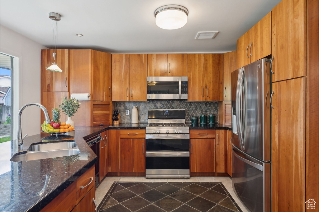 Kitchen with appliances with stainless steel finishes, a sink, brown cabinets, tasteful backsplash, and dark stone counters