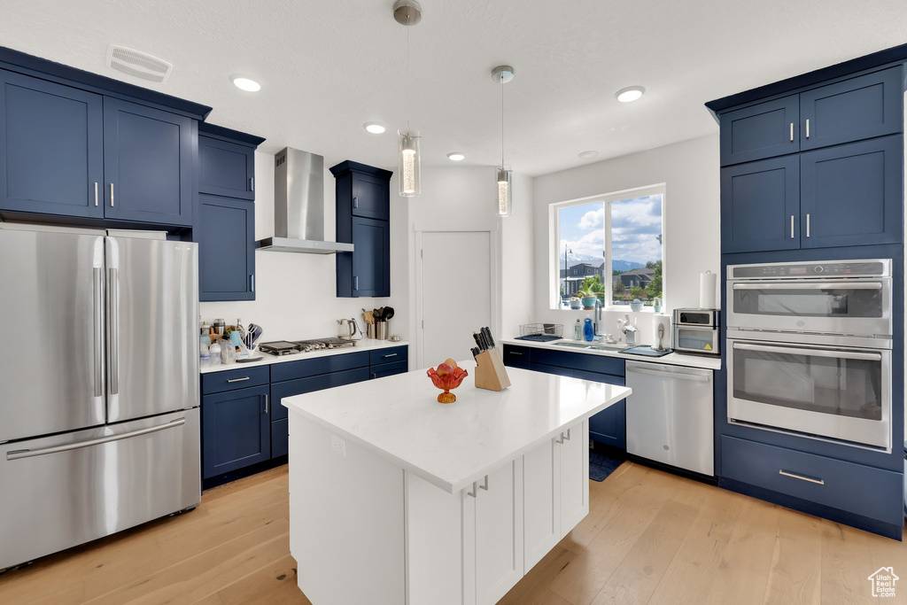 Kitchen featuring stainless steel appliances, wall chimney range hood, blue cabinets, light countertops, and light wood finished floors