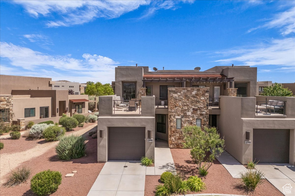 Pueblo revival-style home with a balcony, stucco siding, driveway, stone siding, and a garage