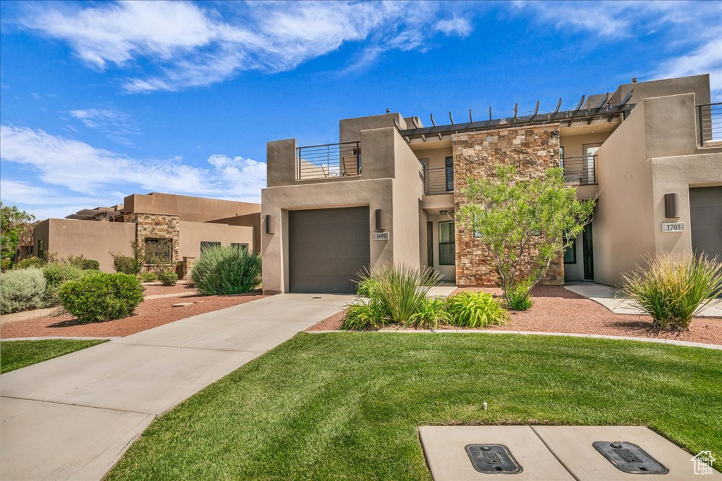 View of front facade featuring a balcony, stone siding, driveway, stucco siding, and a front yard