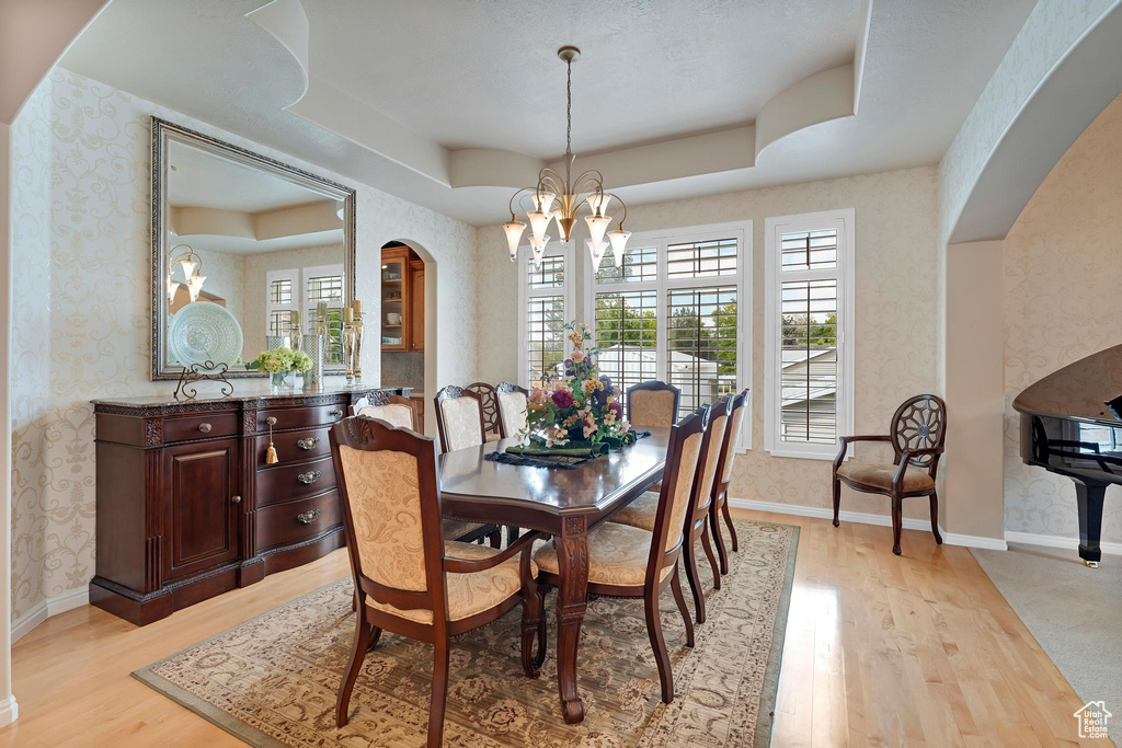 Dining space featuring wallpapered walls, a chandelier, arched walkways, a raised ceiling, and light wood-type flooring