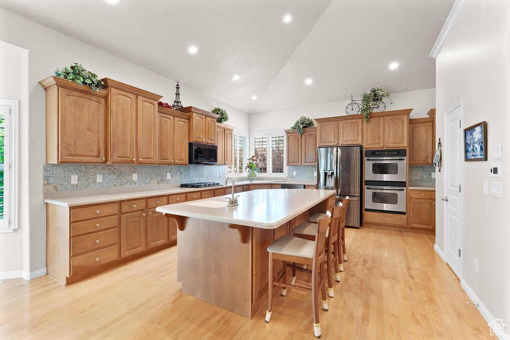 Kitchen with stainless steel appliances, a kitchen island with sink, light countertops, light wood-style floors, and vaulted ceiling