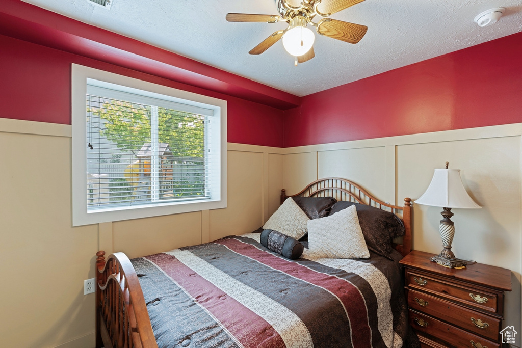 Bedroom featuring ceiling fan, wainscoting, a textured ceiling, and a decorative wall