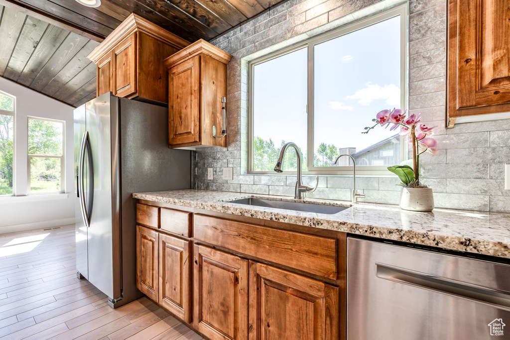 Kitchen with appliances with stainless steel finishes, wood ceiling, a sink, light stone counters, and brown cabinetry