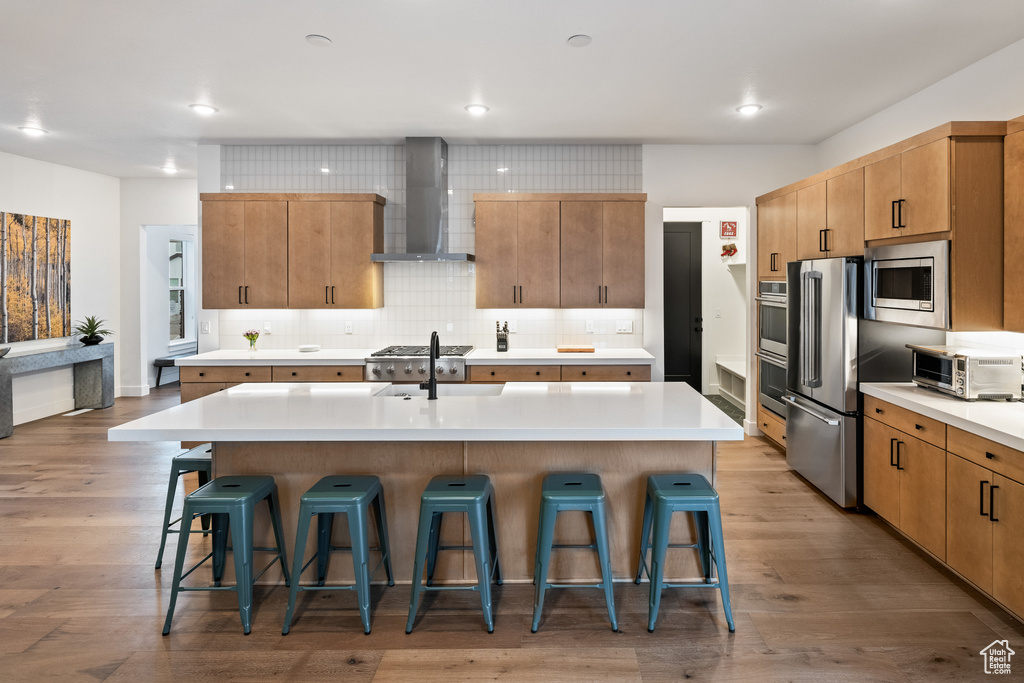 Kitchen with wall chimney exhaust hood, appliances with stainless steel finishes, a kitchen bar, an island with sink, and recessed lighting