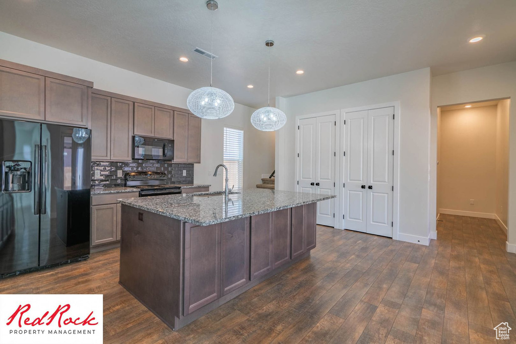 Kitchen featuring black appliances, dark wood-style flooring, light stone counters, an island with sink, and baseboards