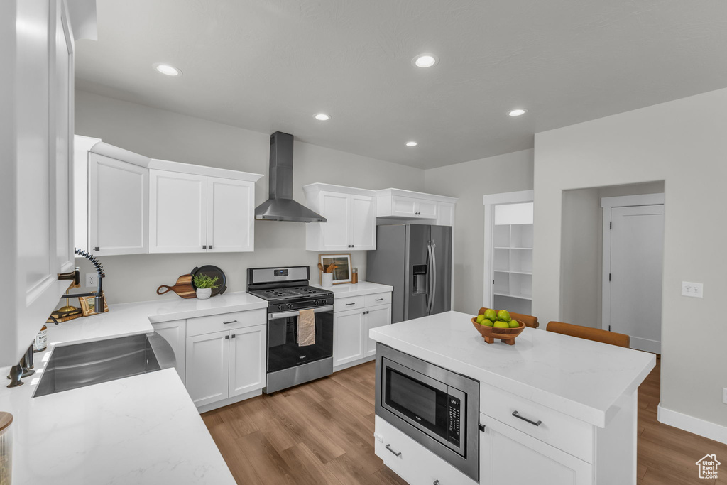 Kitchen featuring wall chimney range hood, appliances with stainless steel finishes, wood finished floors, white cabinets, and recessed lighting
