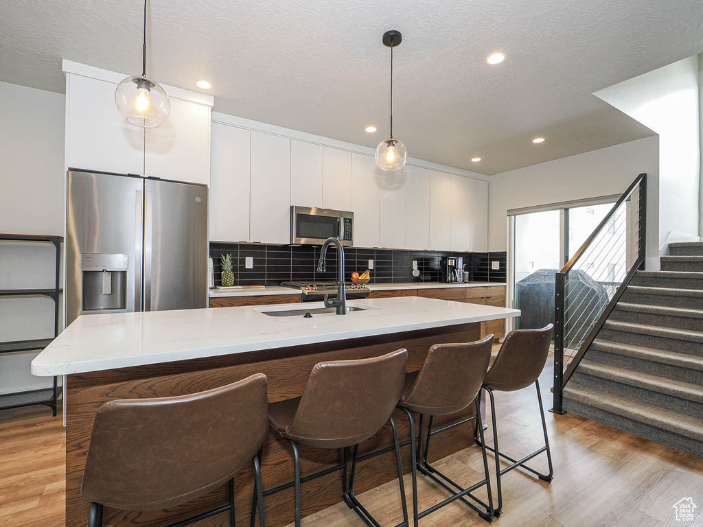 Kitchen with appliances with stainless steel finishes, a sink, white cabinets, a center island with sink, and light wood-type flooring