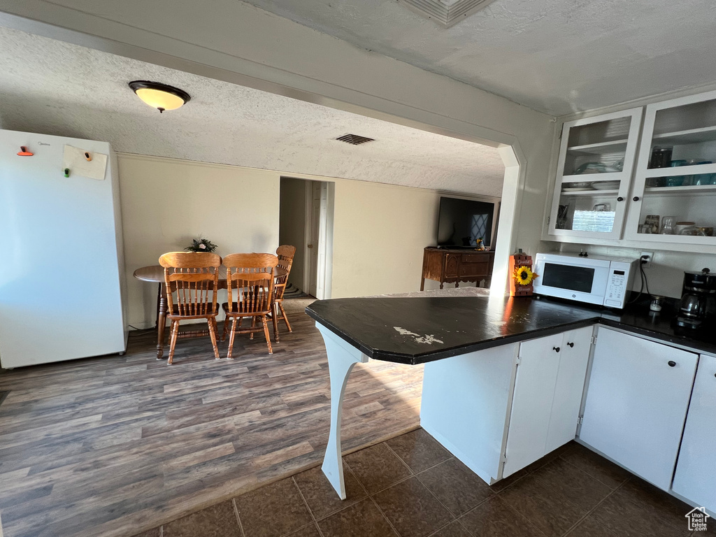 Kitchen with white appliances, dark countertops, a textured ceiling, a peninsula, and glass insert cabinets