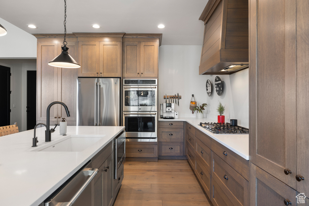 Kitchen featuring appliances with stainless steel finishes, a sink, wall chimney range hood, light countertops, and light wood-type flooring