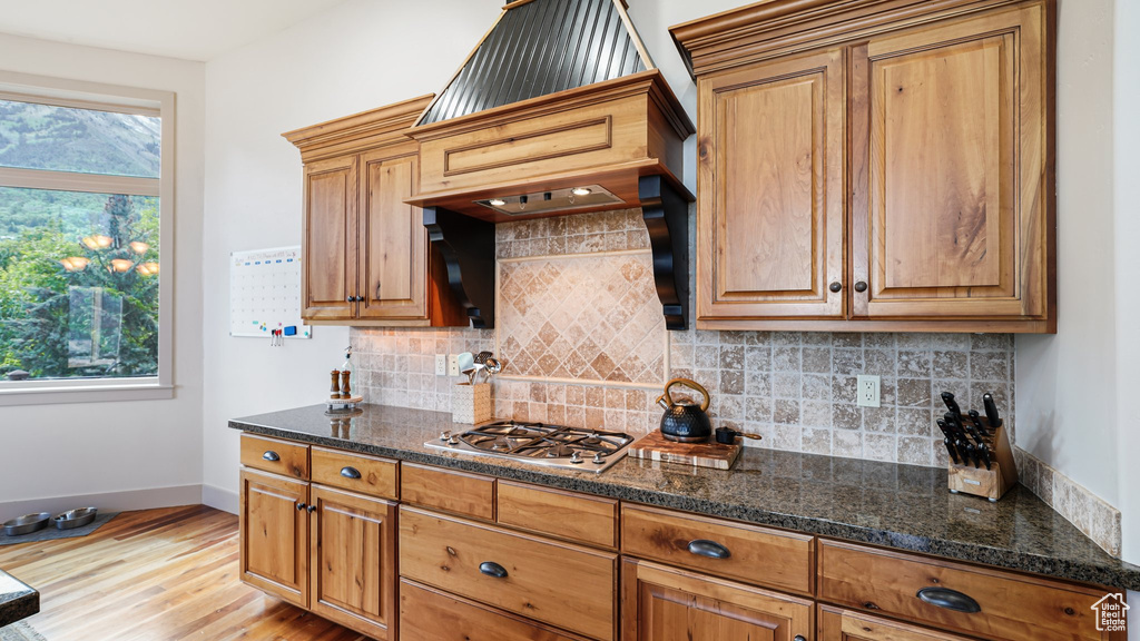 Kitchen with light wood-type flooring, baseboards, stainless steel gas stovetop, backsplash, and dark stone countertops