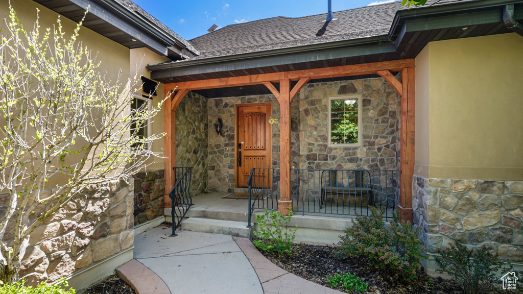 View of exterior entry featuring stone siding, stucco siding, covered porch, and a shingled roof