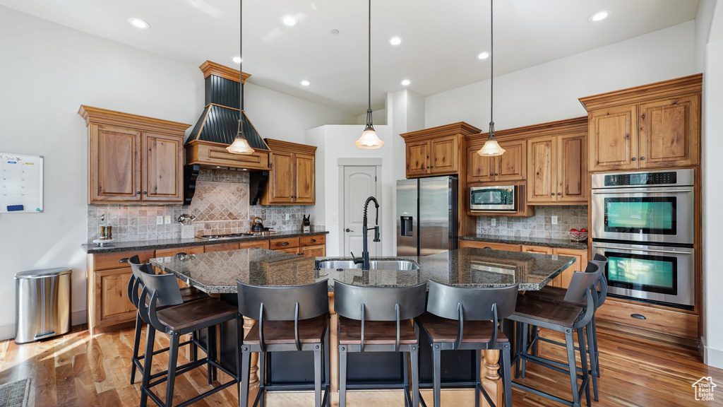 Kitchen with stainless steel appliances, a sink, backsplash, a center island with sink, and brown cabinetry