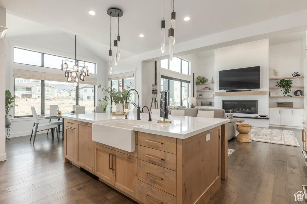 Kitchen with a sink, light brown cabinetry, a glass covered fireplace, hanging light fixtures, and a center island with sink