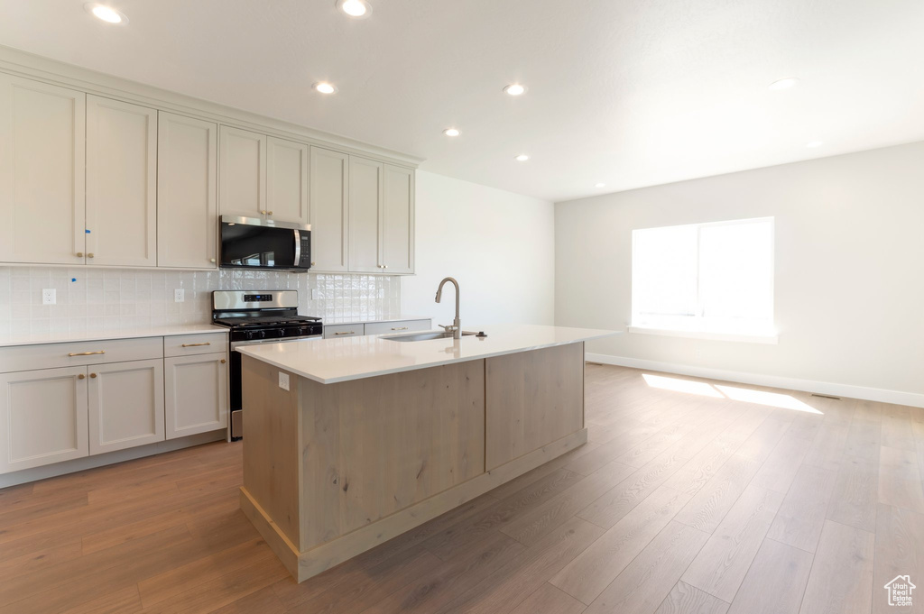 Kitchen featuring stainless steel appliances, a sink, decorative backsplash, a kitchen island with sink, and light wood-type flooring