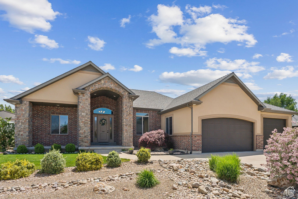 View of front facade featuring brick siding, an attached garage, stucco siding, driveway, and roof with shingles