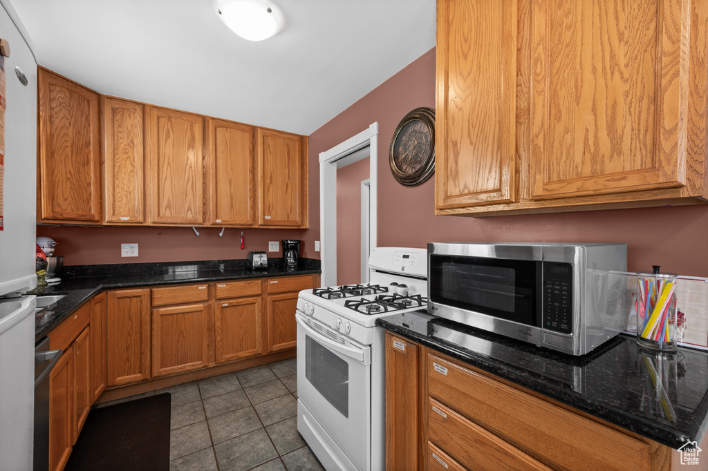 Kitchen with white appliances, tile patterned floors, dark stone countertops, and brown cabinetry