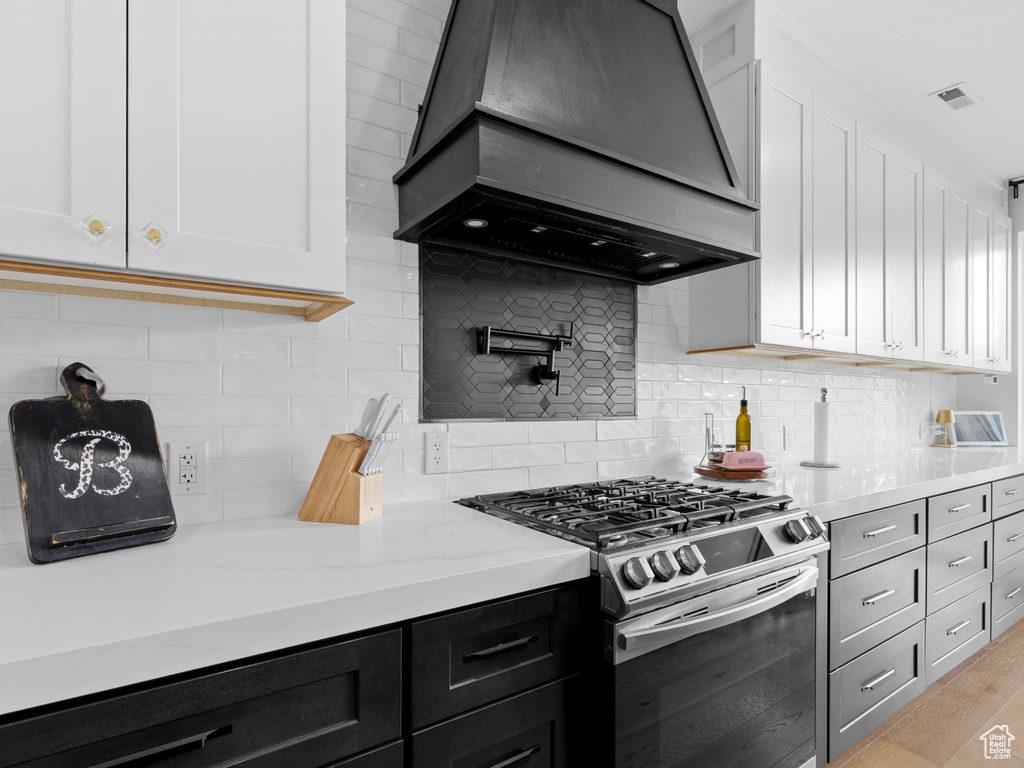 Kitchen featuring custom exhaust hood, stainless steel gas range, white cabinetry, light stone counters, and tasteful backsplash