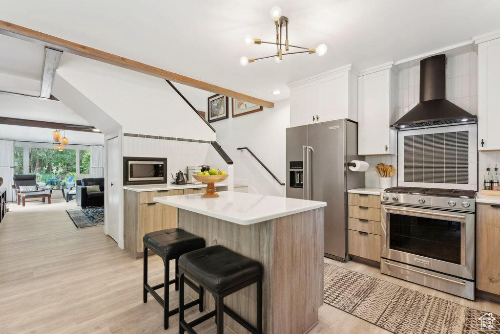 Kitchen featuring stainless steel appliances, wall chimney exhaust hood, a chandelier, light wood-style floors, and beam ceiling