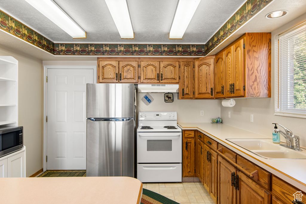 Kitchen with appliances with stainless steel finishes, a sink, under cabinet range hood, brown cabinetry, and light countertops