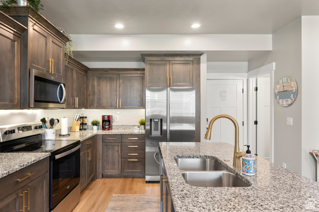 Kitchen with appliances with stainless steel finishes, a sink, light wood finished floors, dark brown cabinets, and light stone countertops