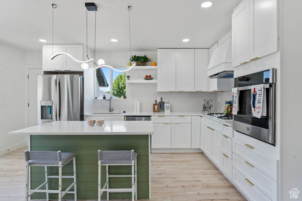 Kitchen featuring appliances with stainless steel finishes, custom range hood, a sink, light wood finished floors, and white cabinetry