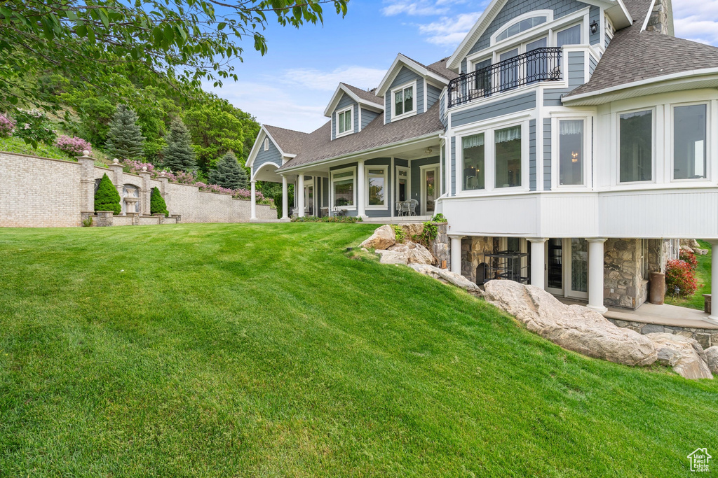 Rear view of property featuring a balcony, a shingled roof, stone siding, and a lawn