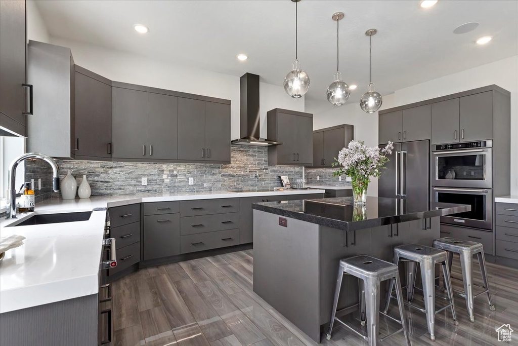 Kitchen with gray cabinetry, appliances with stainless steel finishes, a sink, wall chimney range hood, and a kitchen bar