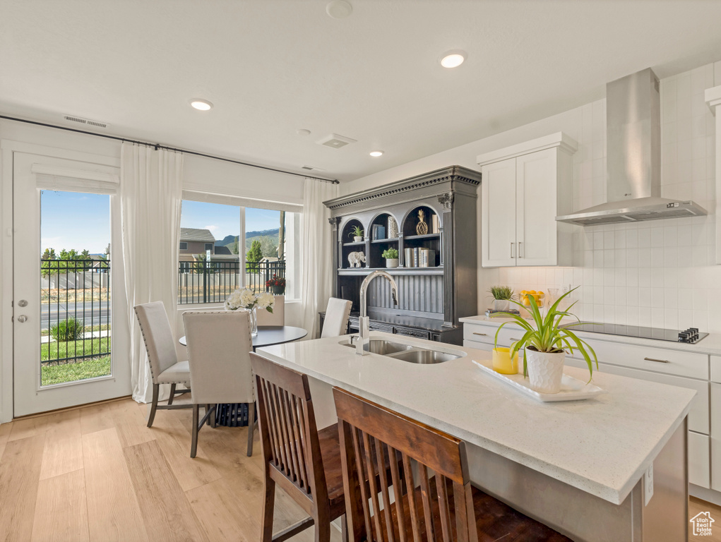 Kitchen with a sink, wall chimney range hood, a kitchen island with sink, black electric cooktop, and light wood-style flooring