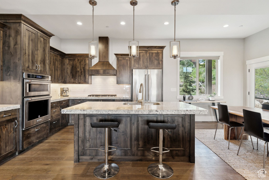 Kitchen with custom exhaust hood, appliances with stainless steel finishes, dark wood finished floors, tasteful backsplash, and a kitchen island with sink