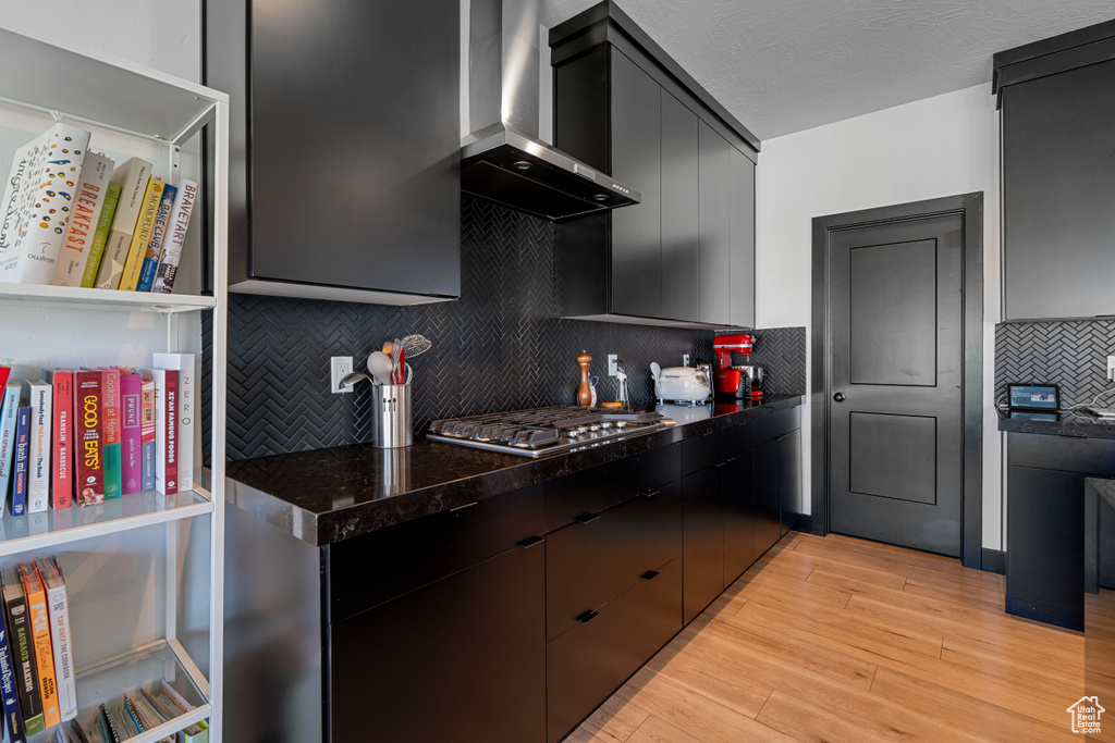 Kitchen featuring wall chimney exhaust hood, stainless steel gas cooktop, backsplash, dark cabinetry, and light wood-type flooring