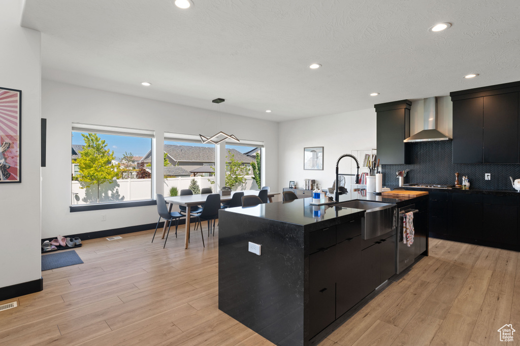 Kitchen with wall chimney exhaust hood, dark cabinetry, a sink, a center island with sink, and decorative backsplash