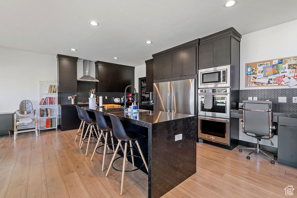 Kitchen with appliances with stainless steel finishes, wall chimney exhaust hood, a sink, light wood-style floors, and a kitchen breakfast bar