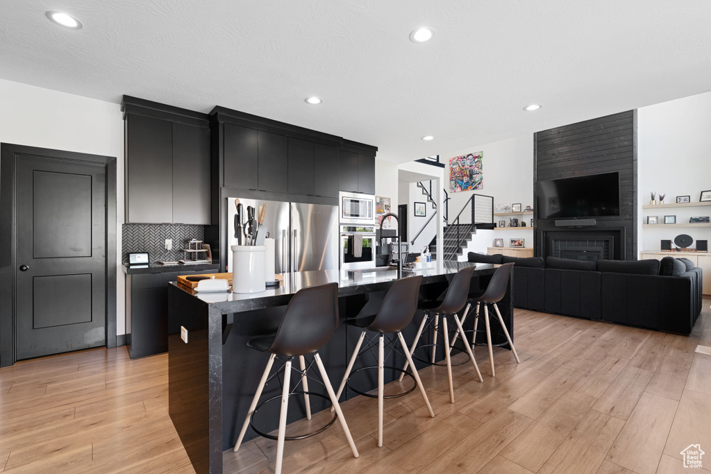 Kitchen with stainless steel appliances, dark cabinetry, a kitchen breakfast bar, an island with sink, and light wood-type flooring