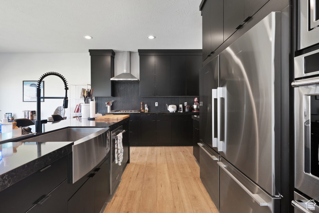 Kitchen featuring stainless steel appliances, dark cabinets, wall chimney exhaust hood, a sink, and backsplash