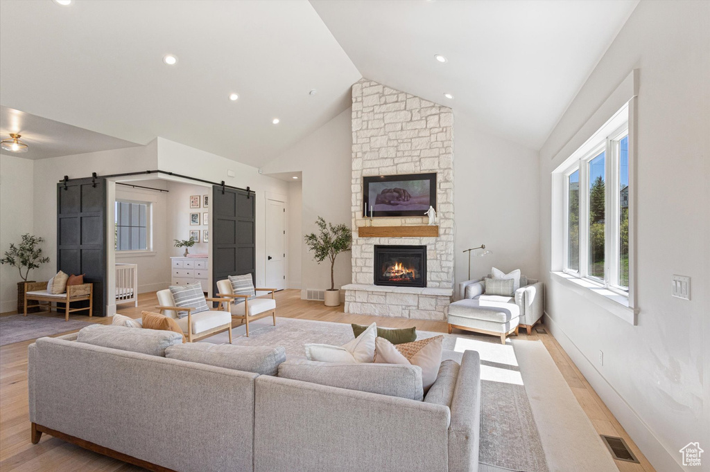 Living room featuring a barn door, light wood-type flooring, high vaulted ceiling, a stone fireplace, and baseboards