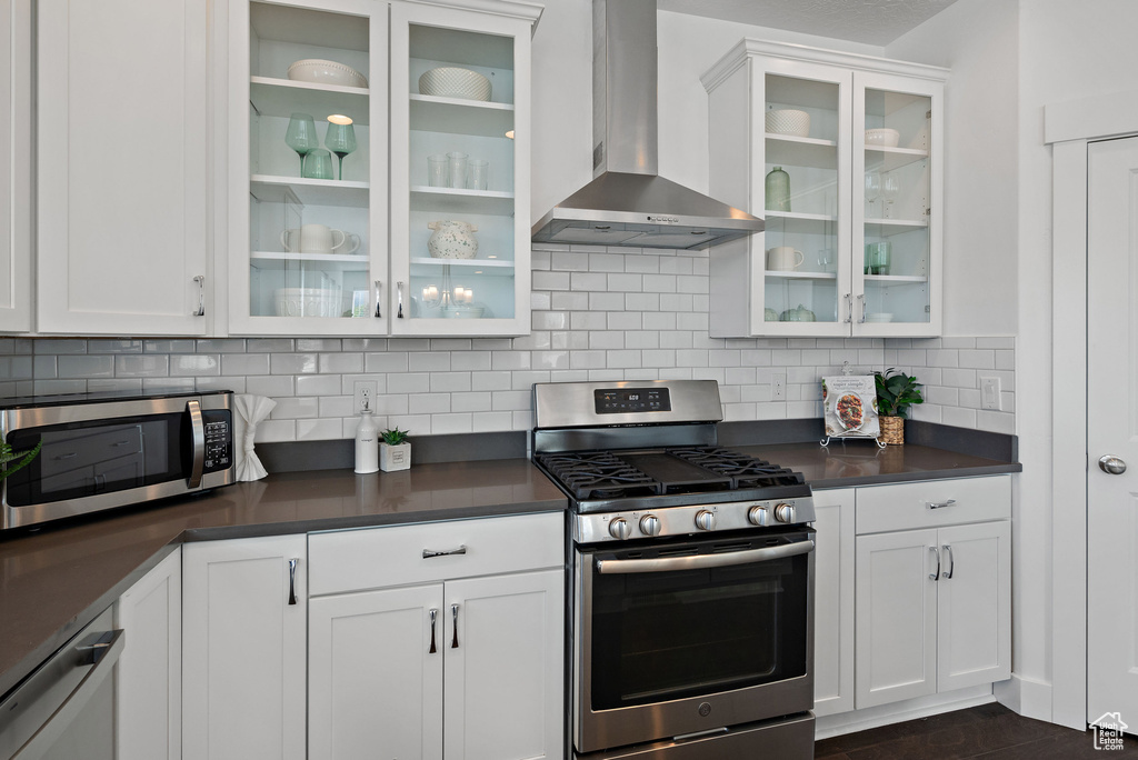 Kitchen with wall chimney range hood, stainless steel appliances, dark countertops, and white cabinetry
