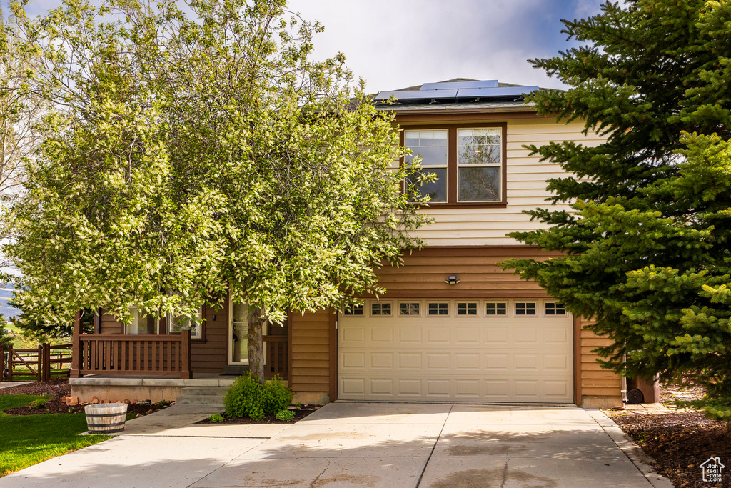 View of property hidden behind natural elements featuring an attached garage, concrete driveway, solar panels, and covered porch