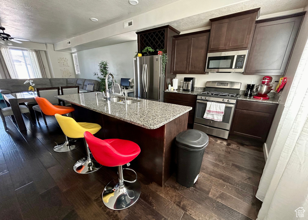 Kitchen featuring appliances with stainless steel finishes, a sink, dark wood finished floors, a kitchen bar, and a kitchen island with sink