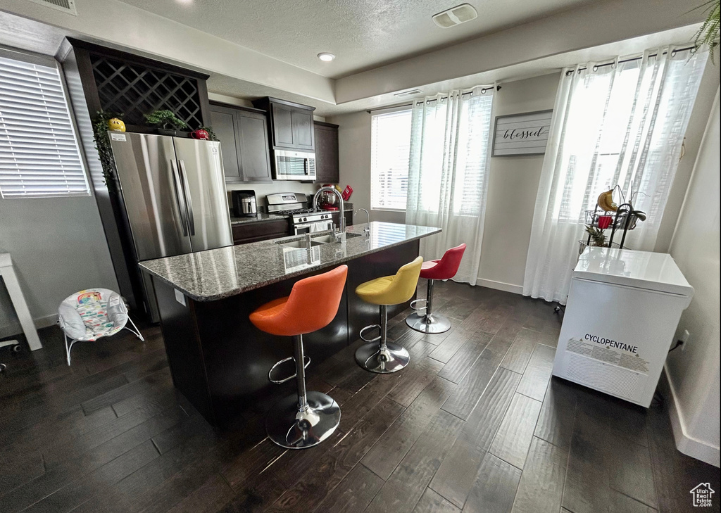 Kitchen featuring stainless steel appliances, dark wood finished floors, a kitchen island with sink, stone counters, and baseboards