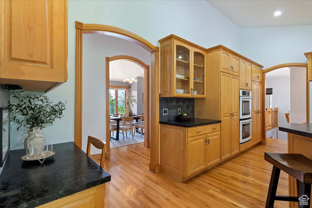 Kitchen featuring arched walkways, light wood-style flooring, double oven, glass insert cabinets, and tasteful backsplash