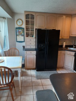 Kitchen featuring black fridge with ice dispenser, a sink, light tile patterned floors, dark countertops, and glass insert cabinets
