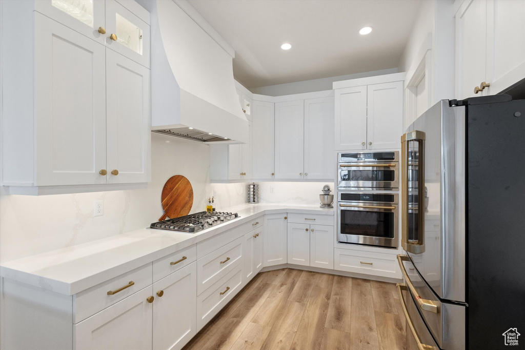 Kitchen featuring appliances with stainless steel finishes, premium range hood, white cabinets, light wood finished floors, and recessed lighting