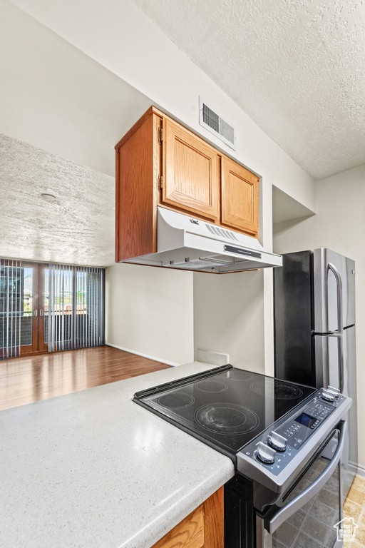 Kitchen with stainless steel electric stove, under cabinet range hood, a textured ceiling, light countertops, and brown cabinetry