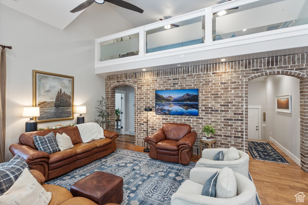 Living room with arched walkways, a ceiling fan, wood finished floors, high vaulted ceiling, and brick wall