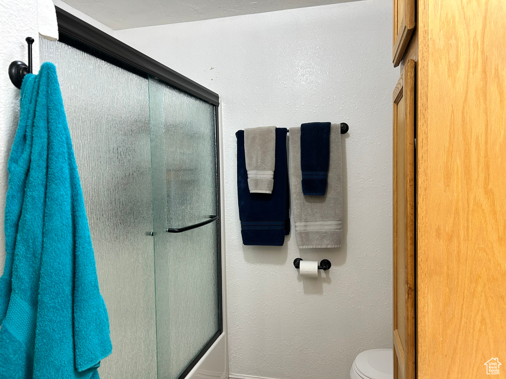 Full bathroom with toilet, bath / shower combo with glass door, and a textured wall