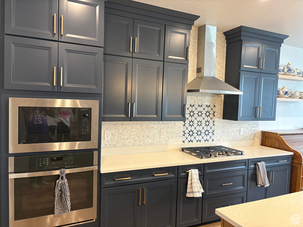 Kitchen featuring appliances with stainless steel finishes, wall chimney range hood, light countertops, decorative backsplash, and open shelves
