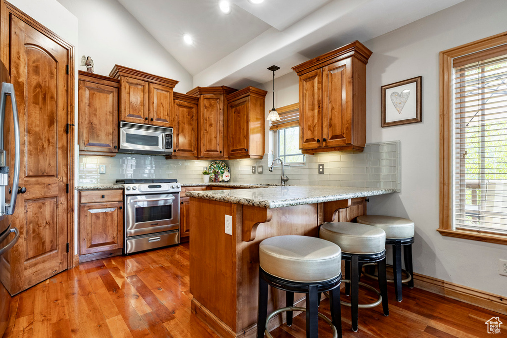 Kitchen with stainless steel appliances, a peninsula, dark wood finished floors, brown cabinetry, and vaulted ceiling